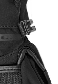 Detalle de una mochila de tela negra que muestra una superficie texturada, una hebilla de plástico negro y un sutil logotipo en el lateral. Presenta acentos de material plegado.