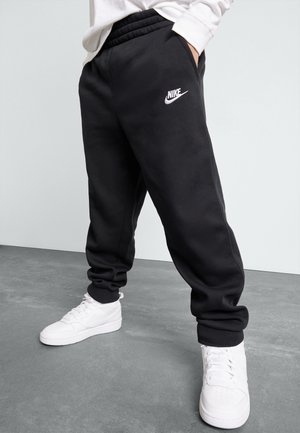 Persoon draagt zwarte Nike sweatpants met logo op de dij en witte sneakers, staand op een grijze vloer tegen een lichte achtergrond.