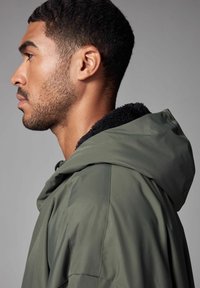 Olivgrüne Kapuzenjacke mit einem schwarzen, flauschigen Kragen. Mit glattem Äußeren und lockerer Passform, seitlich dargestellt.