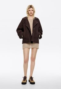 Bruine suède jas met grote kraag, over een lichte, gestructureerde beige trui en een doorzichtige beige rok. Gecombineerd met tan kleurige veterschoenen met zwarte zolen.