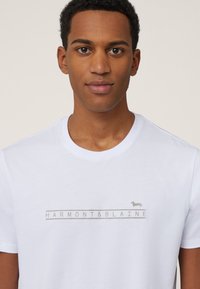 White cotton t-shirt featuring a light gray logo "HARMONT & BLAINE" with a small dachshund graphic centered below the text. Simple crew neckline.