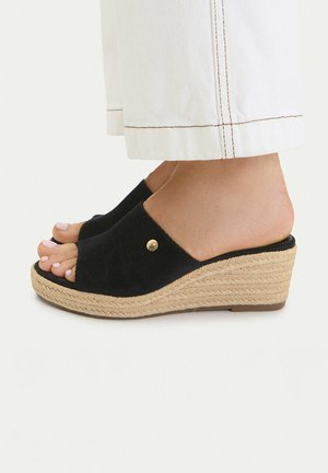 Füße, die schwarze offene Keilsandalen mit geflochtenen Strohsohlen tragen, kombiniert mit weißen Hosen, die über den Knöcheln gekürzt sind.