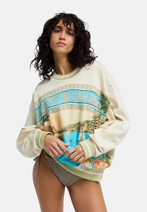 Vrouw met krullend haar draagt een oversized sweatshirt met de tekst "Sunsets in Paradise" en gestreepte bikinibroekjes, poserend tegen een witte achtergrond.