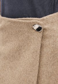 Béžový vlněný oděv s měkkým, strukturovaným povrchem, opatřený kovovým zapínáním na knoflík a čistým, složeným designem.