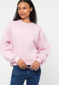 Rózsaszín pamut pulóver kerek nyakkal, ejtett vállakkal és bordázott mandzsettákkal. Az elején fehér színnel nyomtatva van a "TOMMY HILFIGER EST. 1985" felirat.