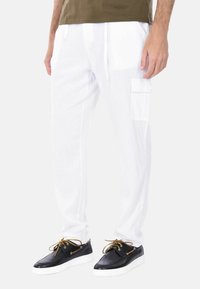Pantaloni cargo bianchi con tasche laterali, realizzati in un tessuto leggero, abbinati a scarpe nere con dettagli dorati e suole bianche.