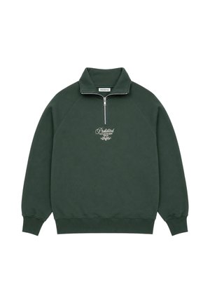 Uomo che indossa una felpa verde scuro con zip corta e scritta bianca, jeans blu e cappello nero che guarda di lato su uno sfondo semplice.