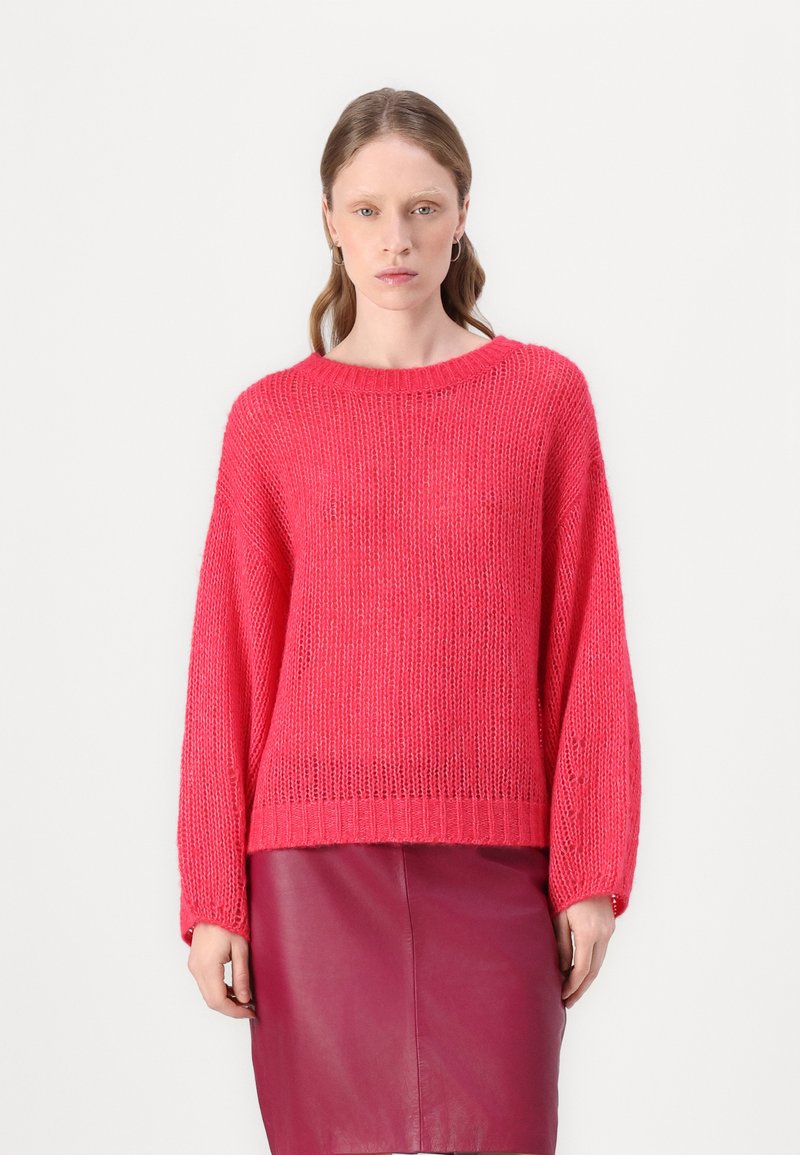 Un pull en tricot rose avec un décolleté large et de longues manches amples, associé à une jupe en cuir bordeaux. Tissu texturé avec une coupe décontractée.
