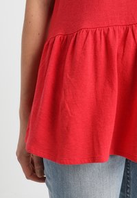 Punainen peplum-toppi pehmeää puuvillaa, jossa on gatheroitu alaosa, hieman löysä siluetti, yhdistettynä vaaleansinsisiin farkkuihin.