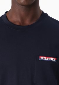 Sötétkék pamut póló kerek nyakkivágással és rövid ujjak. Elöl egy mellzseb található, rajta piros és fehér "HILFIGER" logóval.