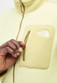 Χέρι με σκουρόχρωμο δέρμα τραβά το φερμουάρ από ένα ανοιχτό κίτρινο φλις με τσέπη στο στήθος και το λογότυπο The North Face.