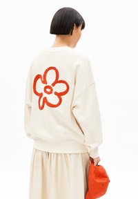 Crème-kleurige oversized sweatshirt met een grote rode bloemenafbeelding op de achterkant. Het model draagt een beige rok en houdt een oranje pet vast.