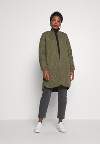 Olivgrön quiltad lång jacka med hög krage, sidfickor och rundad nederkant; parat med svart topp och grå jeans, bärs med vita sneakers.