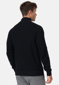 Pull en maille noire avec un col haut, tissu texturé, poignets et ourlet côtelés. La vue de dos montre un design ajusté avec un minimum de détails.