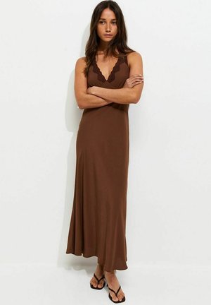 Woman with long dark hair stands with arms crossed wearing a brown sleeveless maxi dress with scalloped neckline and black sandals.