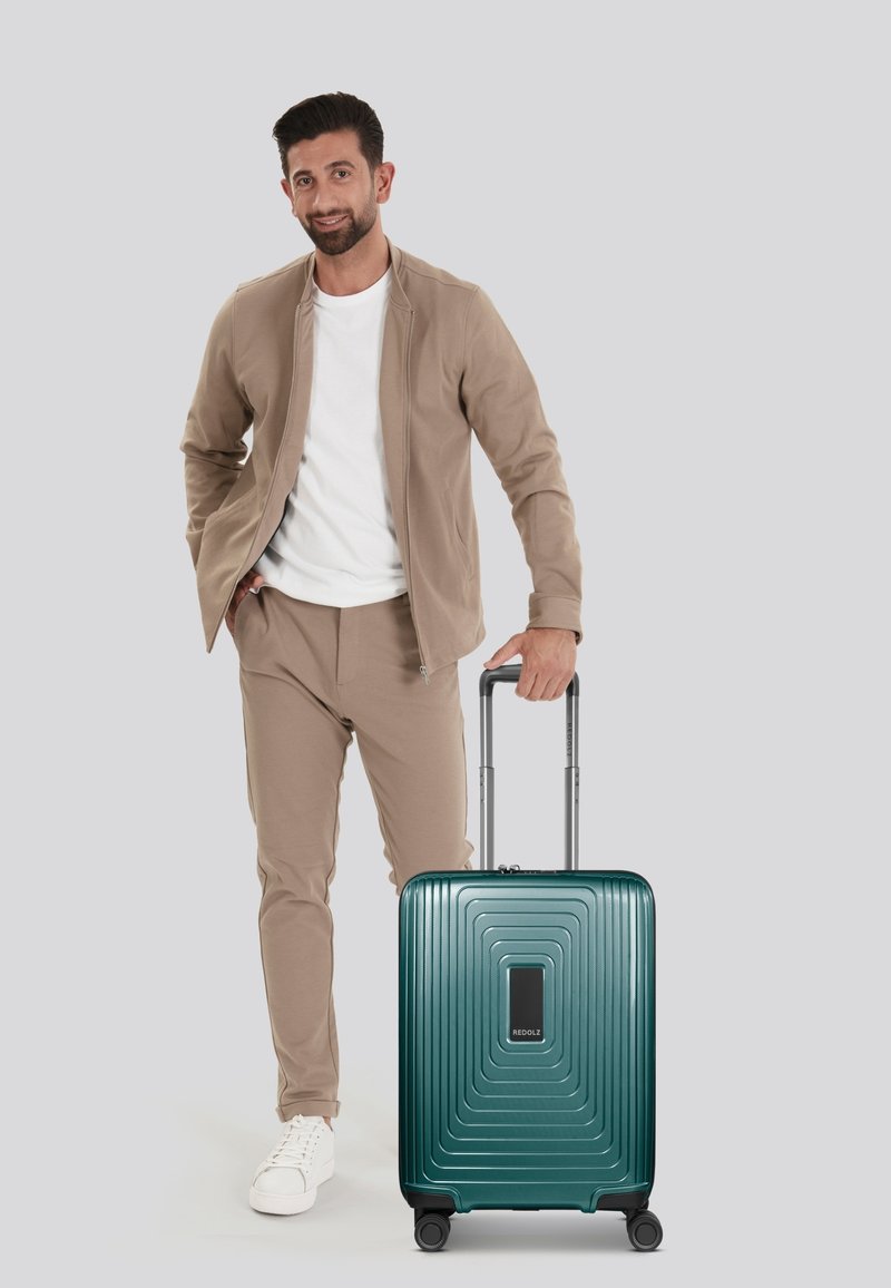 Valigia rigida di un verde scuro con righe orizzontali, dettagli neri, maniglia retrattile e quattro ruote, accanto a un uomo vestito in modo informale.