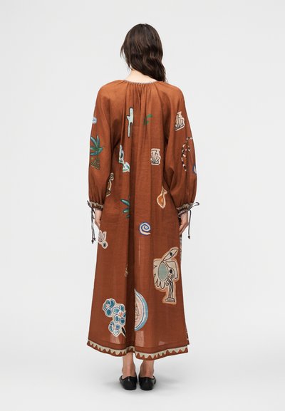 Γυναίκα με μακριά σκούρα μαλλιά, φορώντας ένα φαρδύ, καφέ φόρεμα μέχρι τους αστραγάλους με πολύχρωμα αφηρημένα και βοτανικά σχέδια, όρθια μπροστά σε μια απλή φόντο.