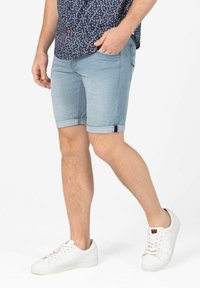 Denim shorts in light blue with rolled hems, featuring a small brand accent on the side. Paired with white sneakers and a patterned shirt.