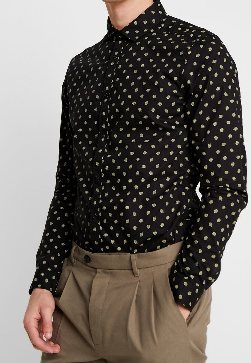 Camicia nera con bottoni e un piccolo motivo a foglie verdi. Presenta un colletto aperto e maniche lunghe, abbinata a pantaloni beige a vita alta.