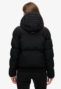 Chaqueta acolchada negra con capucha, con diseño acolchado en forma de cheurón, puños acanalados y un logo en la parte superior de la espalda. Material liso e insonorizado.