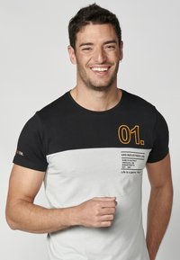 Muška majica s crnim gornjim dijelom i svjetlosivim donjim dijelom. Sadrži otisnutu oznaku i "01." u narančastoj boji. Kratki rukavi, okrugli izrez.