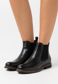 Bottines en cuir noir avec des panneaux élastiques sur les côtés, une pointe ronde et un petit talon carré. Présente des coutures contrastantes en brun le long de la semelle.