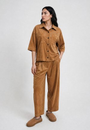 Woman standing with hand in pocket wearing loose brown button-up shirt, matching wide-leg pants, and brown slip-on shoes.