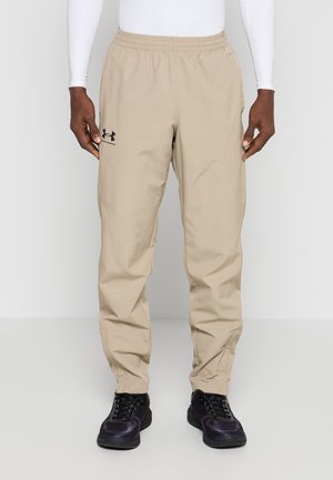 Beżowe spodnie treningowe Under Armour z elastycznym pasem noszone przez osobę w białym topie i czarnych sportowych butach, stojącą na neutralnym tle.