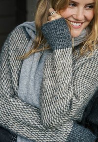 Džemper od pletenine u sivoj i krem boji, s teksturiranim kockastim uzorkom. Dugih rukava s rebrastim manšetama, nošen preko sivog topić.