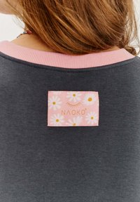 Tmavosivá látka s ružovým ribovaným výstrihom, s obdĺžnikovou etiketou s kvetinovým vzorom a nápisom "NAOKO" zlatým písmom.