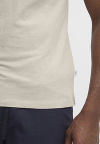 Tricou bej deschis cu o textură moale, având un croi drept și o etichetă mică a mărcii pe partea stângă, aproape de tiv.