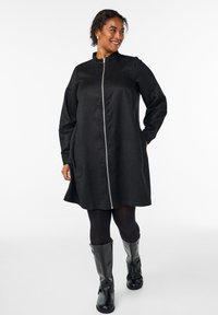 Schwarzes, zip-up Kleid aus glattem Stoff. Lange Ärmel, gerader Schnitt und Seitentaschen. Getragen mit schwarzen Strumpfhosen und knielangen Stiefeln.
