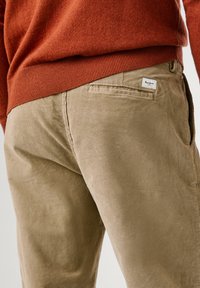 Tan corduroy broek met verticale ribbels, voorzien van een achterzak met een label. Gecombineerd met een donkeroranjeruimte trui.
