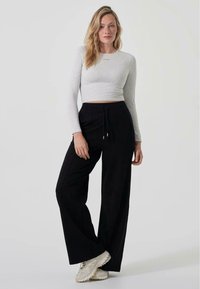 Grå langt ermet cropped topp med tekstdetalj, kombinert med svarte vide bukser med elastisk midje og snøring, iført lette joggesko.