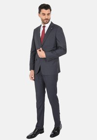 Un abito grigio scuro dal taglio slim, la giacca presenta una tasca singola, abbinata a una camicia bianca e una cravatta rossa. Le scarpe in pelle nera completano il look.
