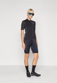 Schwarzes Radtrikot mit kurzen Ärmeln und einem Frontreißverschluss, kombiniert mit schwarzen gepolsterten Shorts. Reflektierende Sonnenbrille und schwarze Radsportschuhe vervollständigen das Outfit.