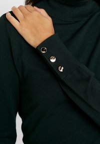 Dunkelgrüner Rollkragenpullover mit gerippter Textur, der drei Akzente mit goldfarbener Knopfdetails am Bund aufweist. Sleek, taillierter Schnitt.