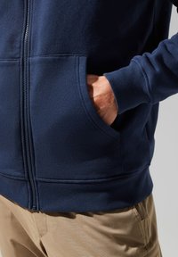 Navy blue zip-up hoodie made of soft fabric, featuring a kangaroo pocket and ribbed cuffs. Visible hand in pocket, neutral-colored pants below.