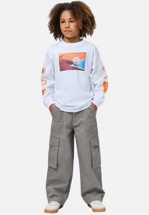 Bambino con capelli ricci, mani nelle tasche, indossa una maglietta bianca a maniche lunghe con stampa di un campo da basket e pantaloni cargo grigi.