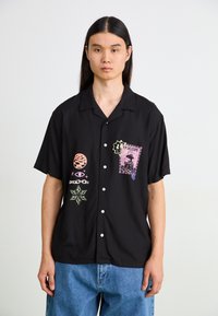 Chemise noire à manches courtes avec des graphiques brodés colorés, y compris des formes géométriques et des motifs, et des boutons blancs à l'avant.