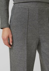 Szare wełniane spodnie o gładkiej fakturze z wysokim stanem, przednimi zakładkami i bocznymi kieszeniami, z ręką opartą na materiale.