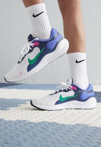 Baskets de sport blanches avec des accents verts et roses, tige en maille, semelle rembourrée et talon violet contrastant. Portées avec des chaussettes blanches de type crew.