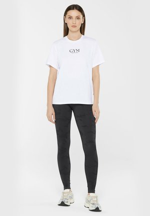 Weißes Baumwoll-T-Shirt mit schwarzem "GYM"-Schriftzug, kombiniert mit schwarzen Camo-leggings und beigen Sportschuhen. Schlichte, lockere Passform.