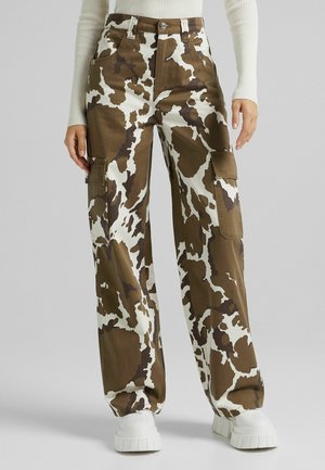 Brązowo-białe spodnie cargo w motyw krowy noszone z białymi butami na grubej podeszwie i kremowym, prążkowanym topem z długim rękawem.