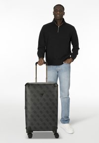 Valigia nera con motivo a fantasia, finitura lucida, angoli opachi, manico telescopico e quattro ruote, mostrata accanto a una persona con abbigliamento casual.