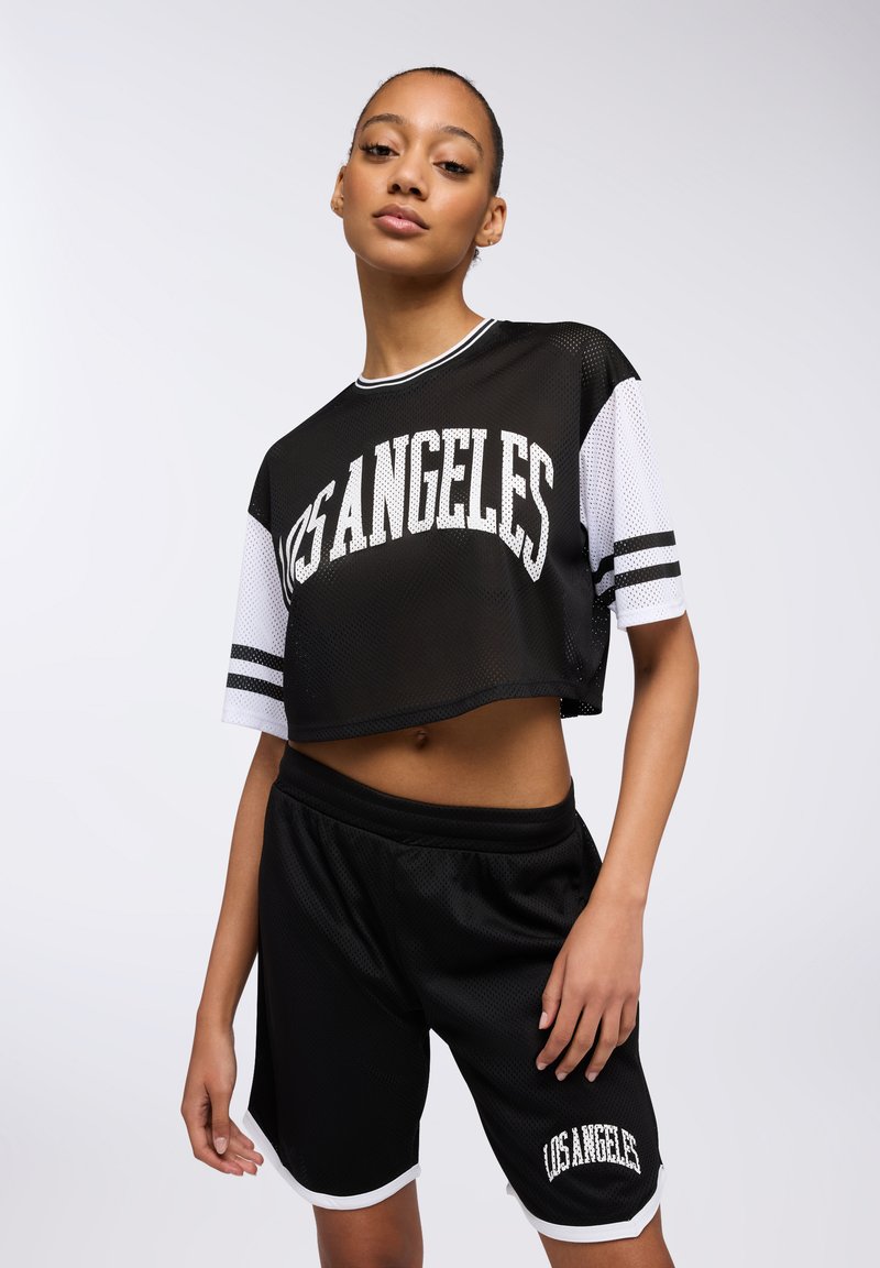 Maglia nera a rete corta con la scritta "LOS ANGELES" stampata, abbinata a pantaloni corti neri con bordatura bianca e logo coordinato, il tutto in stile atletico.