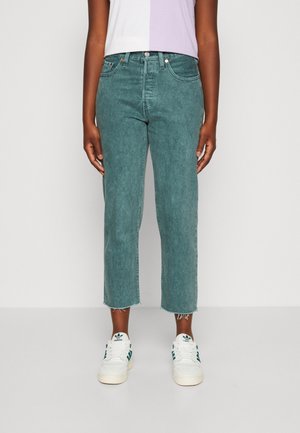 Person trägt verkürzte, ausgewaschene blaue gerade Jeans mit rohen Säumen und weißen Adidas-Sneakern mit grünen Streifen.