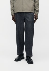 Homme portant un jean large foncé avec des coutures roses, des mocassins en cuir noir et une veste gris clair sur un fond blanc uni.