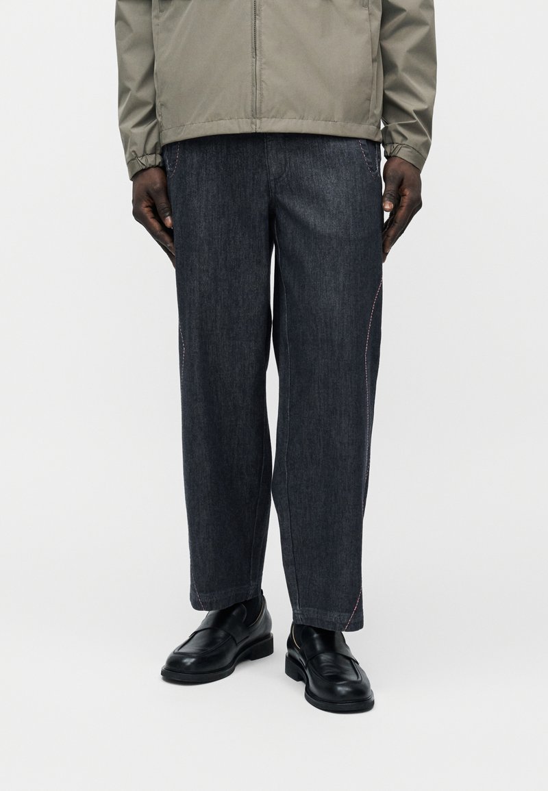 Homme portant un jean large foncé avec des coutures roses, des mocassins en cuir noir et une veste gris clair sur un fond blanc uni.