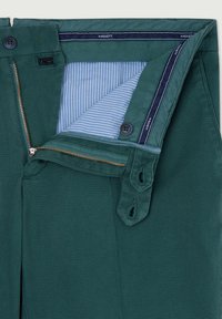 Pantalon en coton vert avec braguette zippée à quincaillerie en laiton, doublure intérieure de taille et de poche à rayures bleues, avec des accents de boutons.